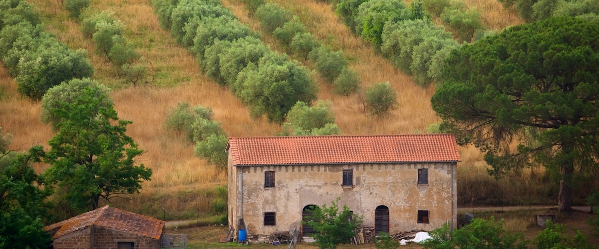 A rustic building with a red-tiled roof surrounded by a hilly landscape with dense green trees.