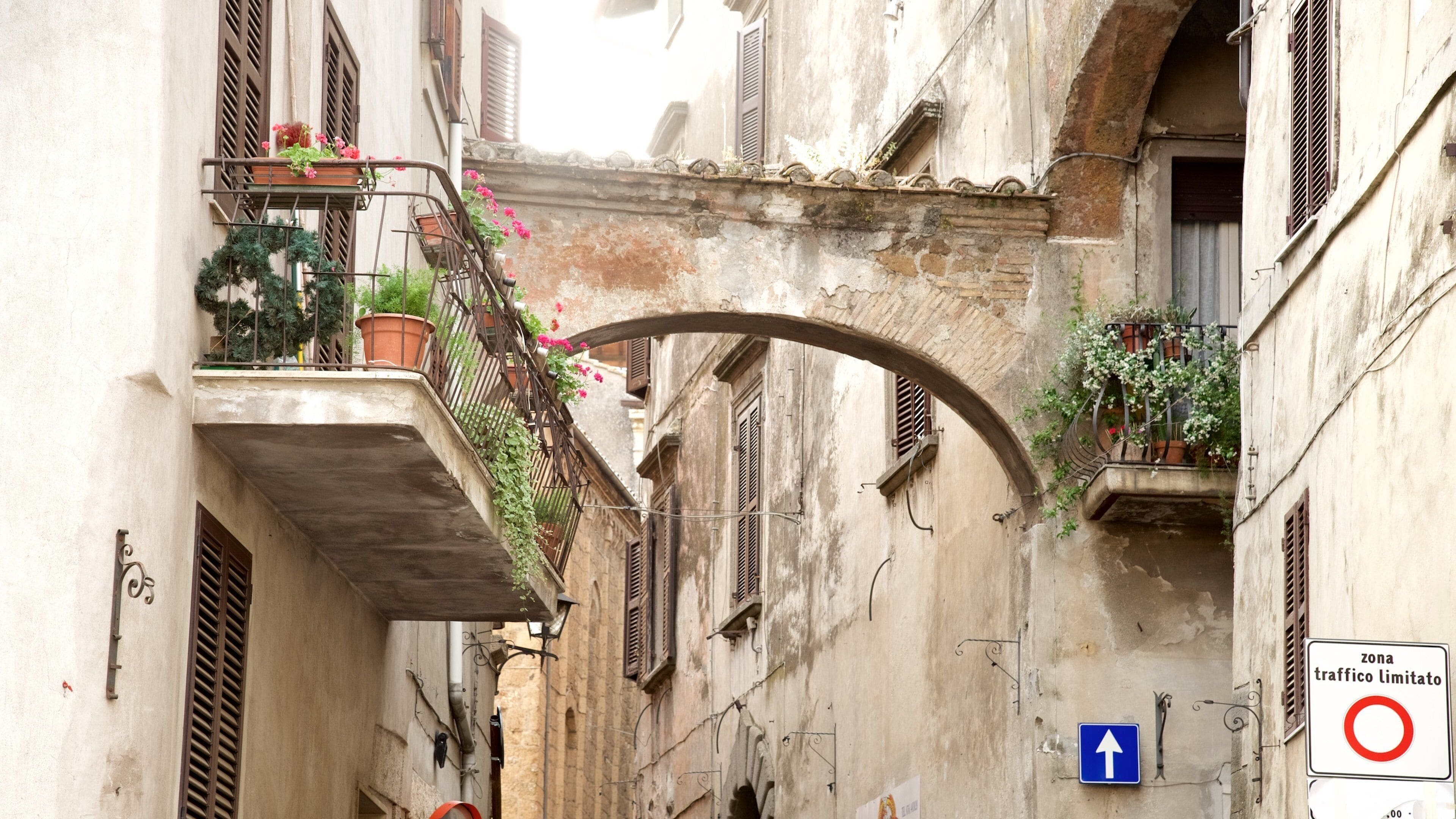 A narrow street with a stone bridge, balconies with potted plants, and a traffic sign indicating a limited traffic zone.
