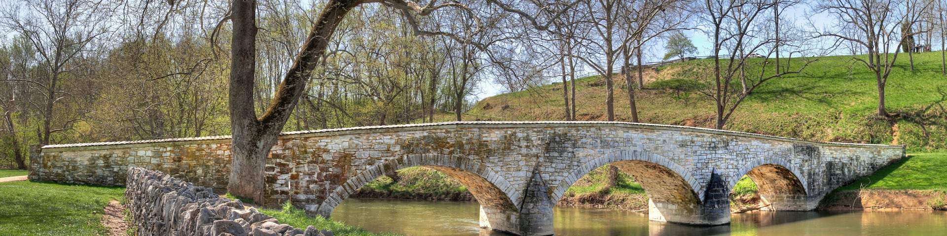 A stone bridge over a calm river, surrounded by trees and grassy hills.