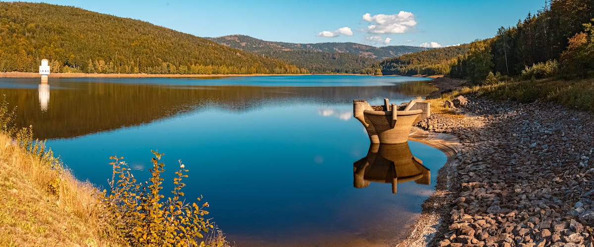 A calm lake with a concrete structure in the center, surrounded by rocky shores and forested hills.