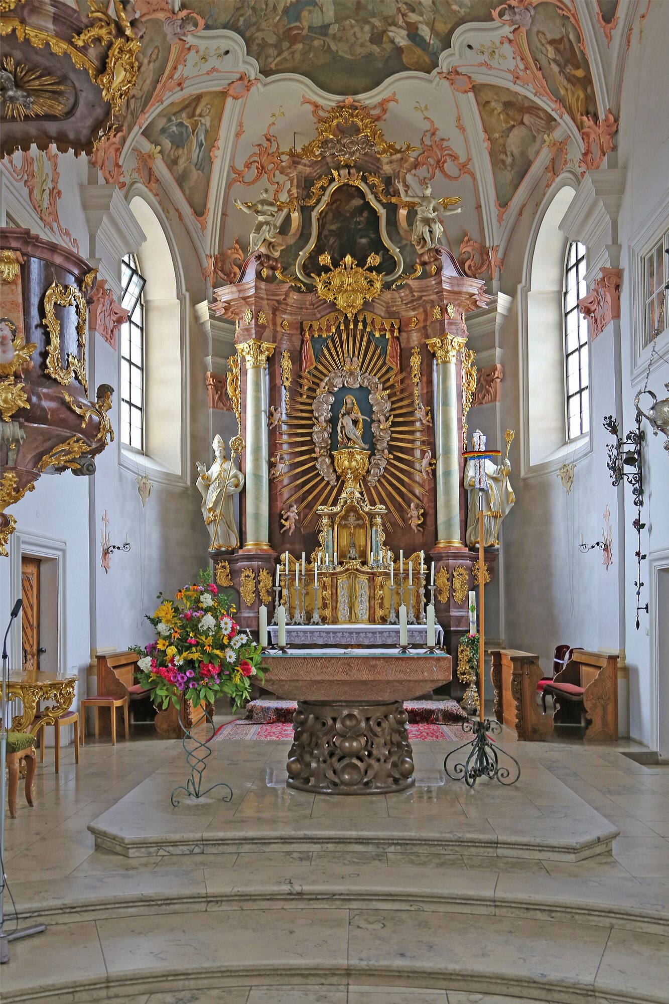Un altar ornamentado con estatuas religiosas y una figura central, rodeado de arreglos florales y velas.