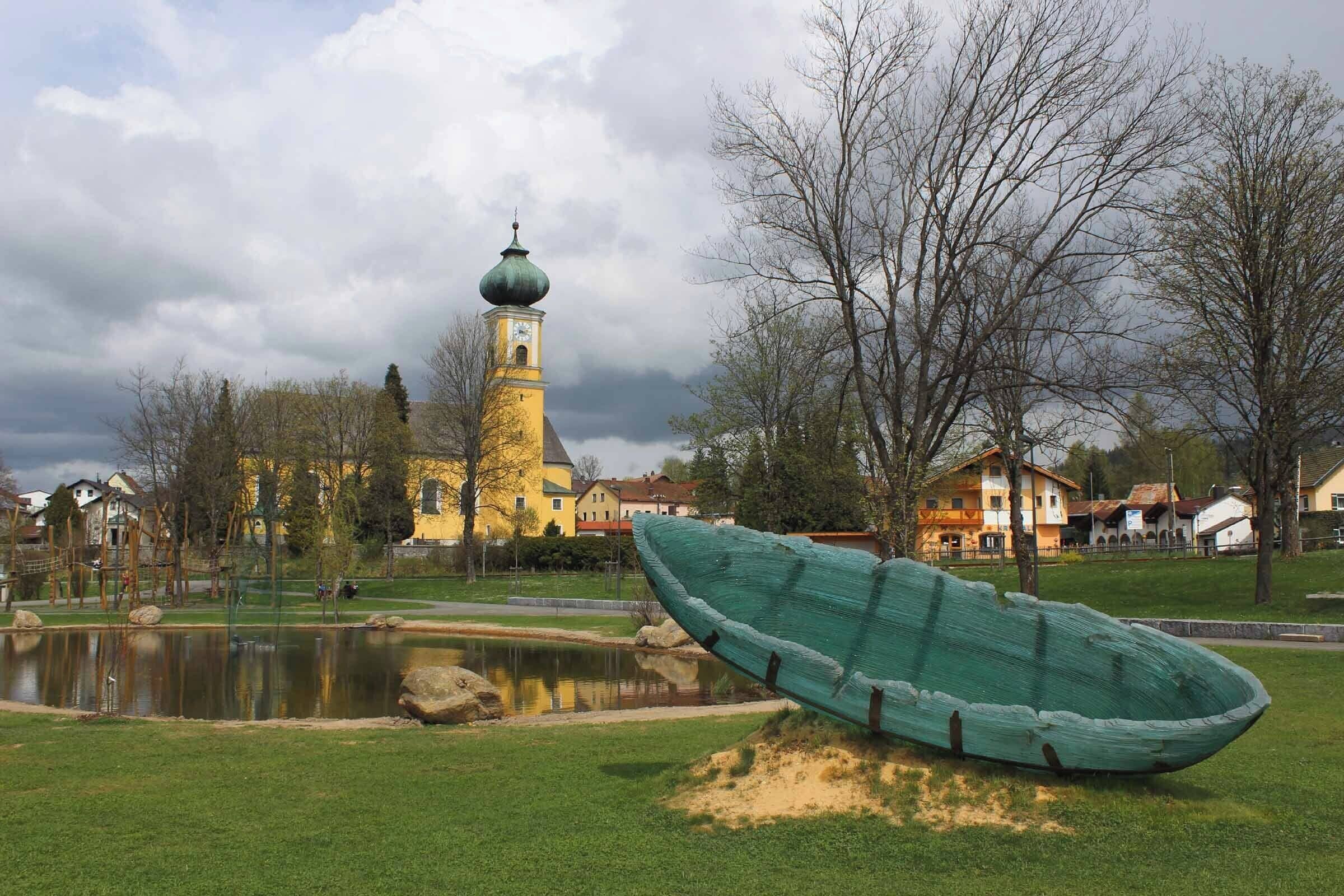 Un bote verde sobre un área cubierta de césped, un estanque y una iglesia con cúpula verde al fondo.