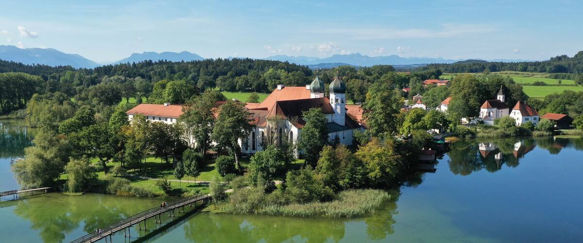 A lakeside village with a church, surrounded by lush greenery and mountains in the background.