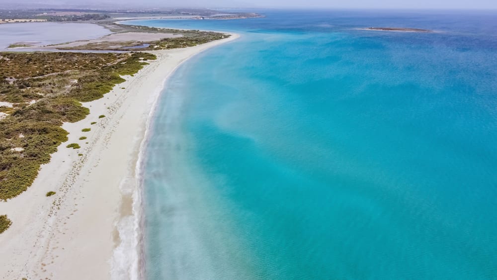 A coastal landscape with a wide sandy beach, clear turquoise waters, and a distant shoreline.