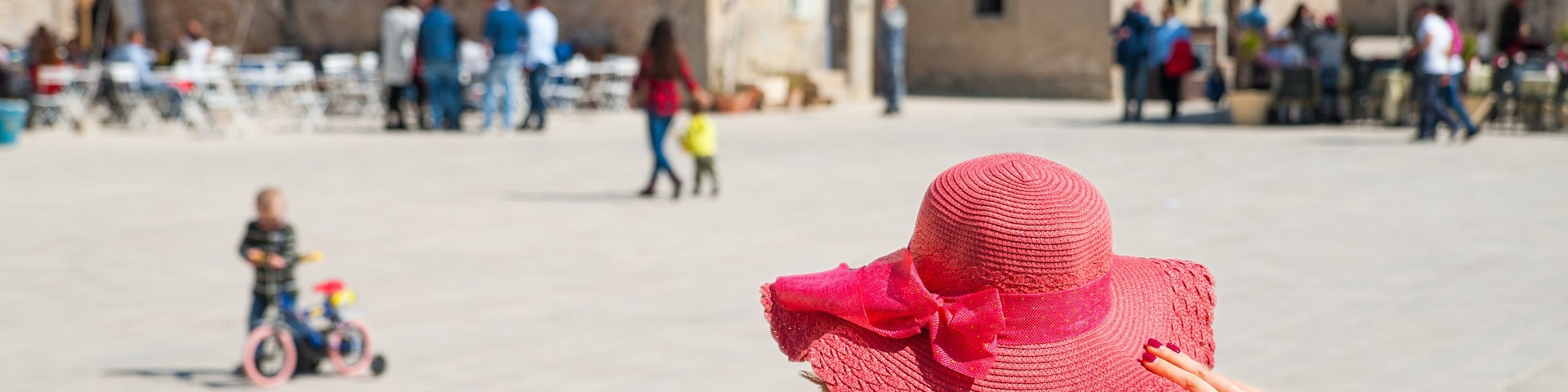 A person wearing a pink hat and denim jacket in a square with stone buildings and outdoor seating.