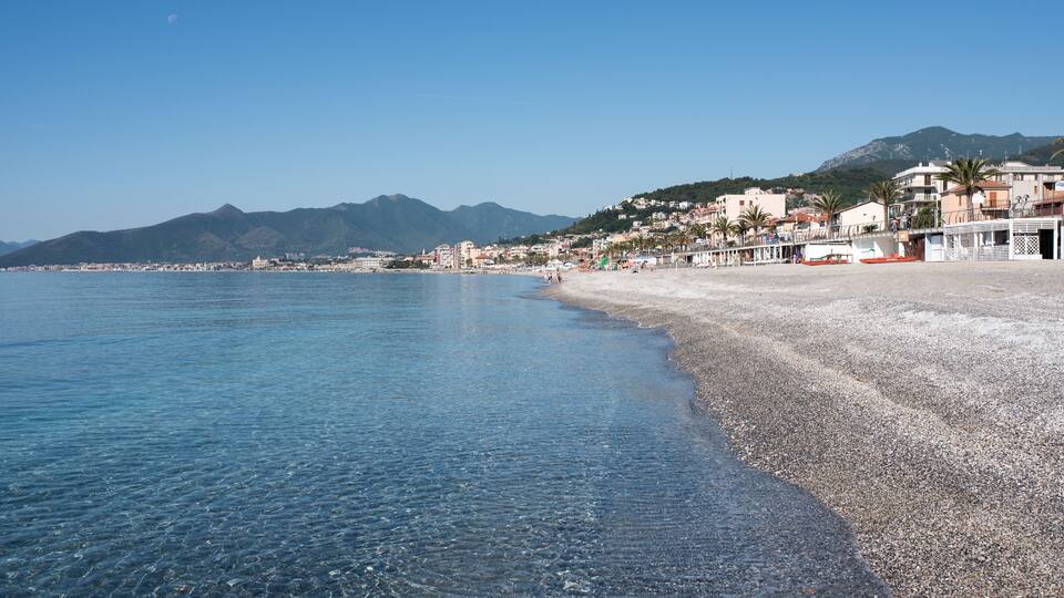 A coastal beach with clear blue water, pebbly shore, and buildings along the coastline.
