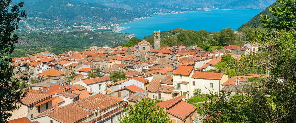 A coastal town with red-roofed buildings, a prominent church with a bell tower, and a backdrop of mountains and a clear blue sea.