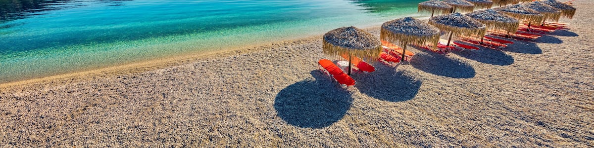 A pebbly beach with clear turquoise water, a row of straw umbrellas, and green hills in the background.
