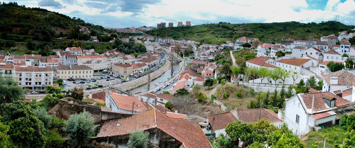 A town with terracotta-roofed buildings, a winding road, and a hill with greenery.