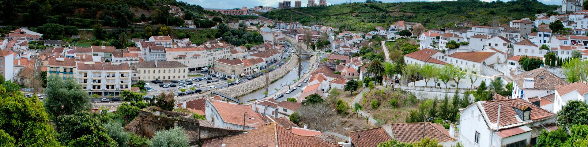 A town with terracotta-roofed buildings, a winding road, and a hill with greenery.