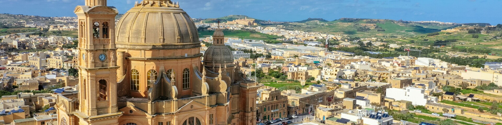 A large domed cathedral with a bell tower, surrounded by a densely packed urban area with flat-roofed buildings and a hilly landscape in the background.