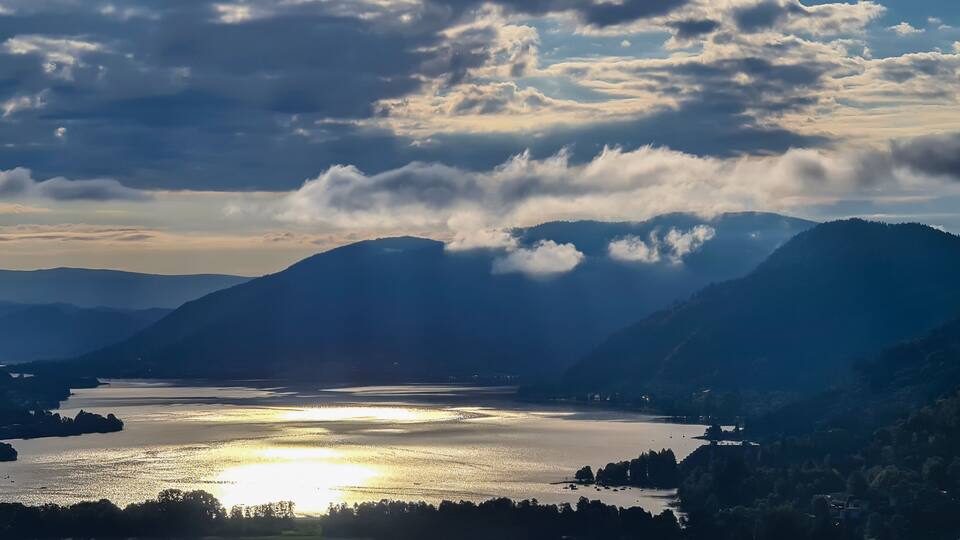 A mountainous landscape with a lake reflecting sunlight, surrounded by hills and a partly cloudy sky.