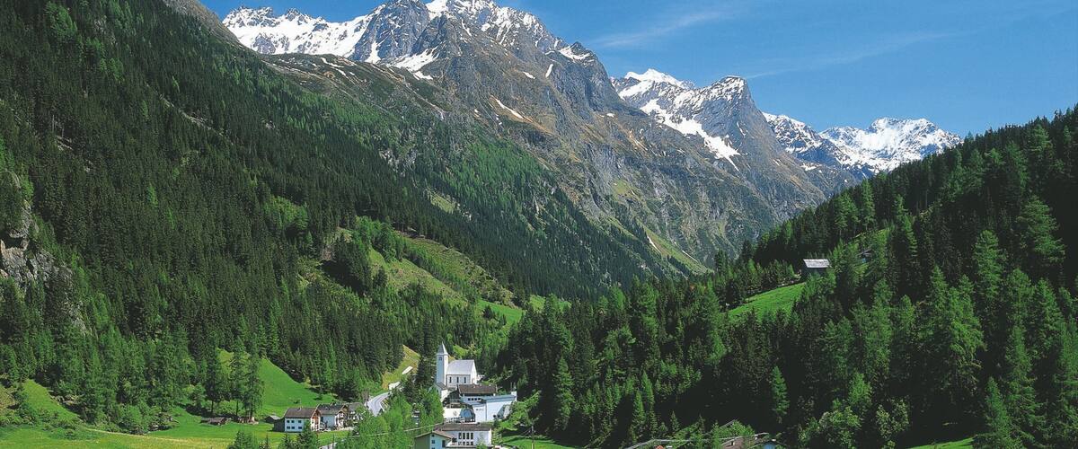 A mountainous landscape with a village nestled in a valley, surrounded by dense forests and snow-capped peaks.