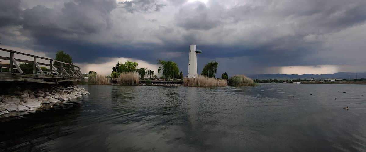 A river with a white tower, a wooden bridge, and a cloudy sky.