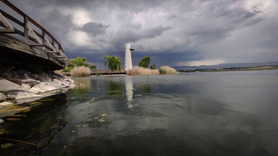 A calm river with a wooden bridge, a tall monument, and a cloudy sky.