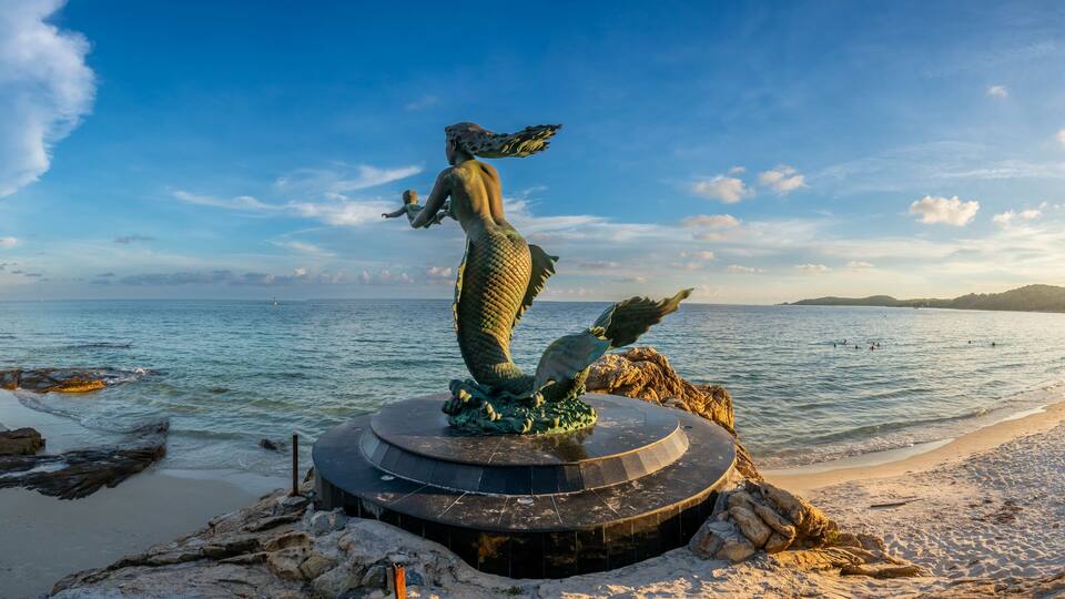 A mermaid statue on a rocky outcrop by a beach with clear blue water and a distant island.