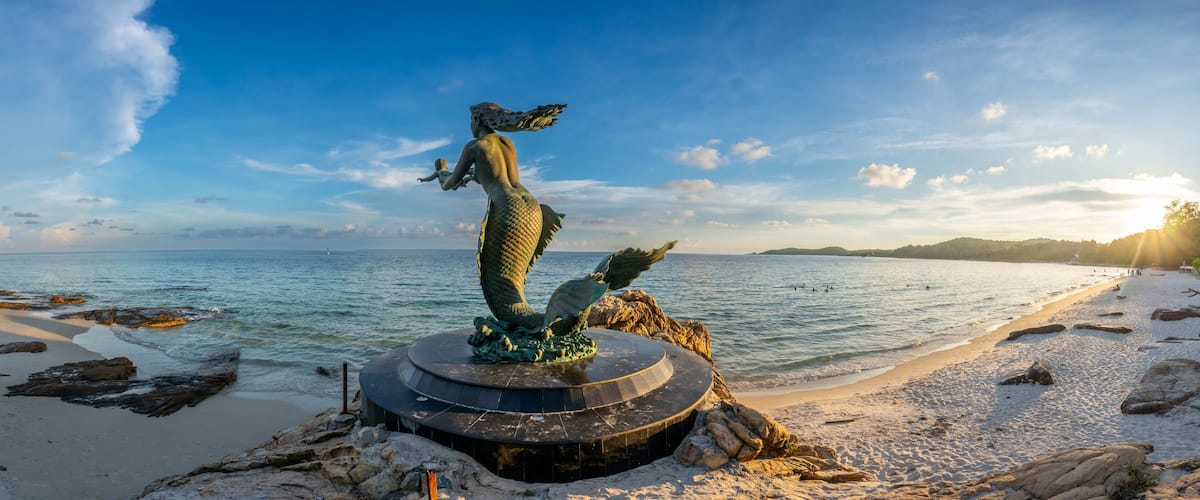 A mermaid statue on a rocky outcrop by a beach with clear blue water and a distant island.