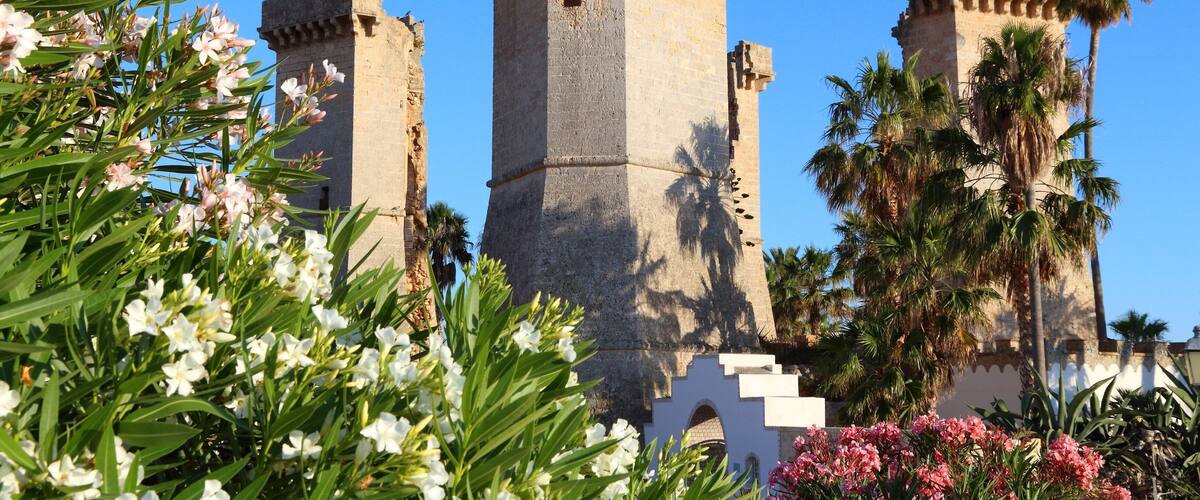 A historic stone tower with a square base and crenellations, surrounded by lush greenery and flowering plants.
