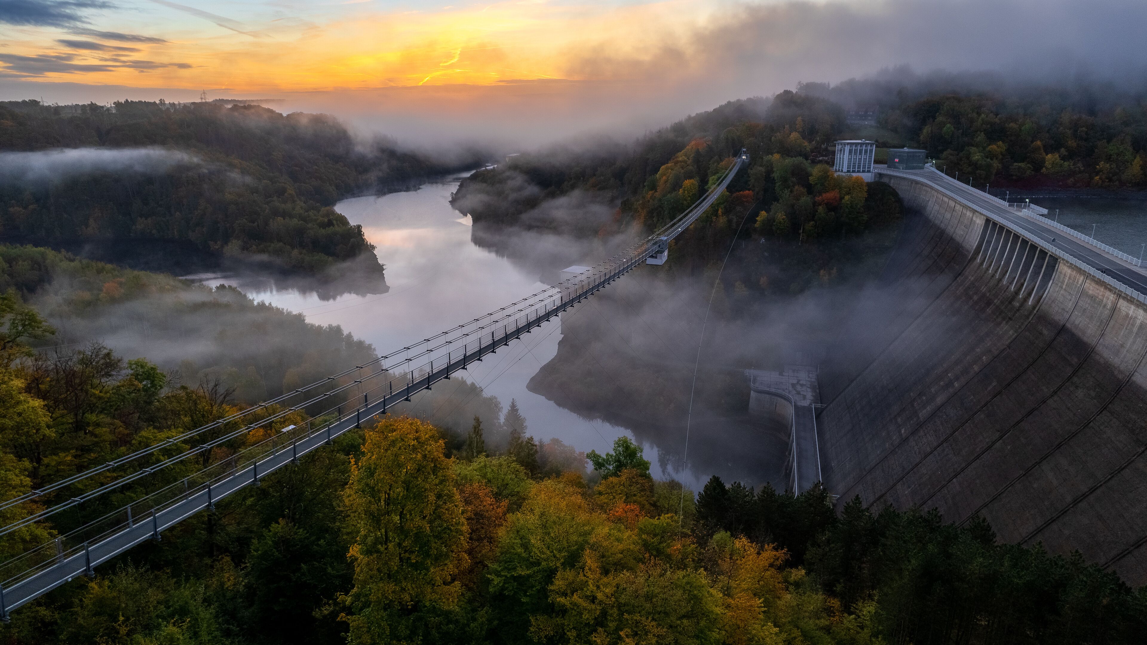 Un pont suspendu enjambant une rivière, entouré de collines boisées et enveloppé de brume.