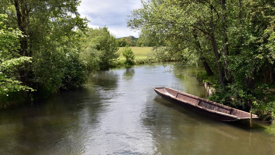 A calm river with a boat docked on the right bank, surrounded by lush greenery and trees.