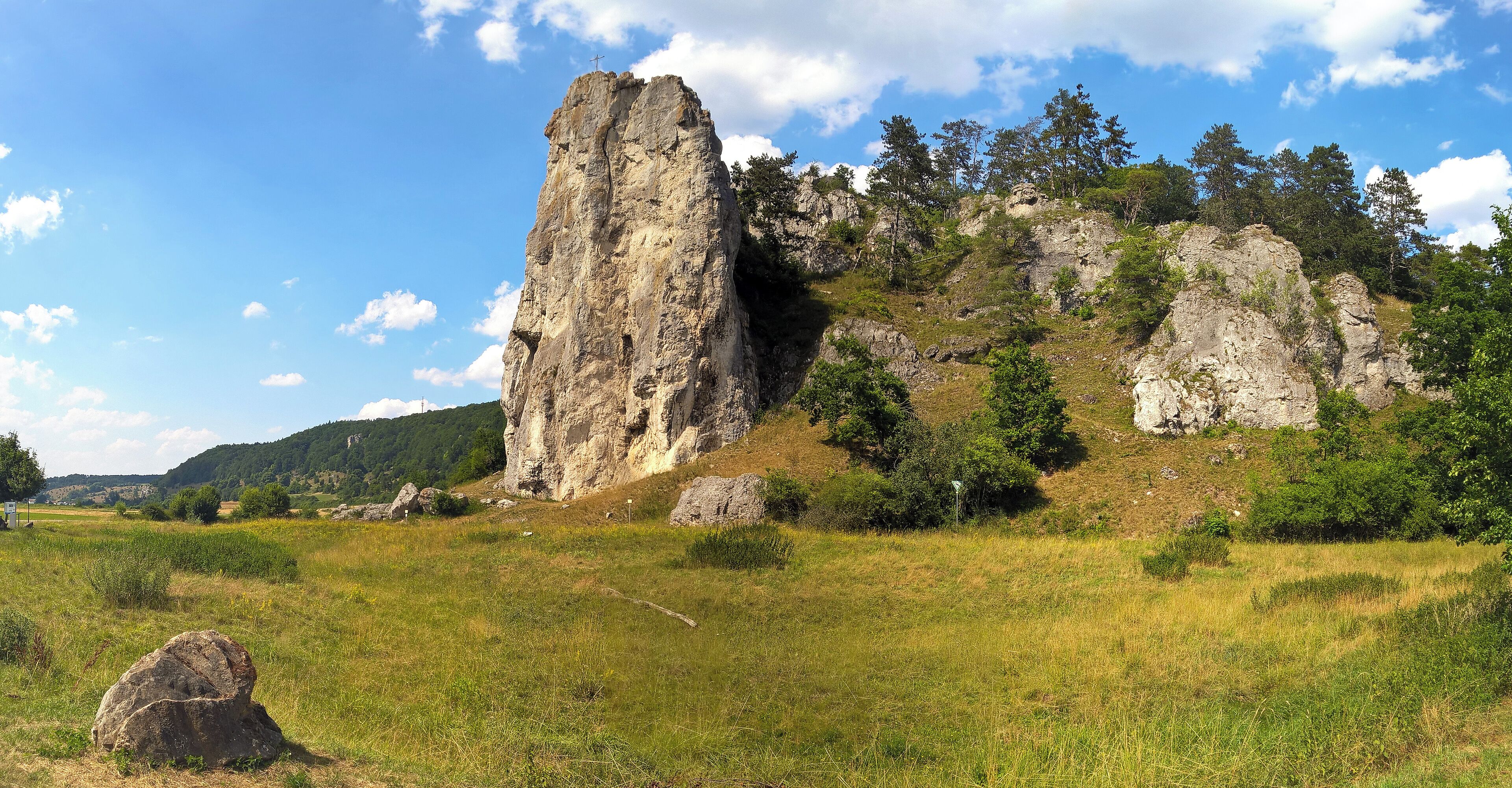 A rocky cliff with a cross at the top, surrounded by greenery and trees.