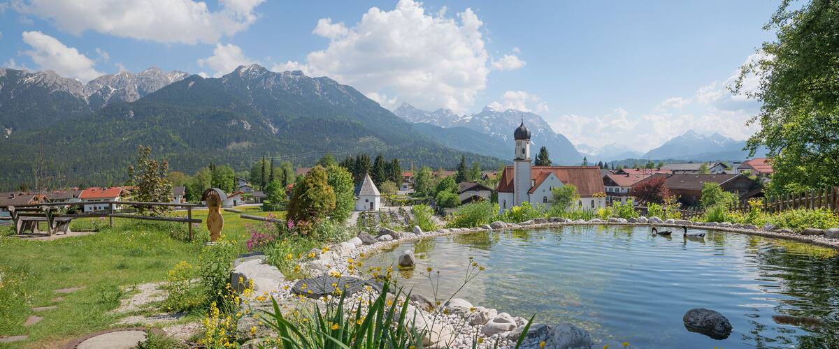 A serene lakeside village with a church, mountains in the background, and a wooden bridge.
