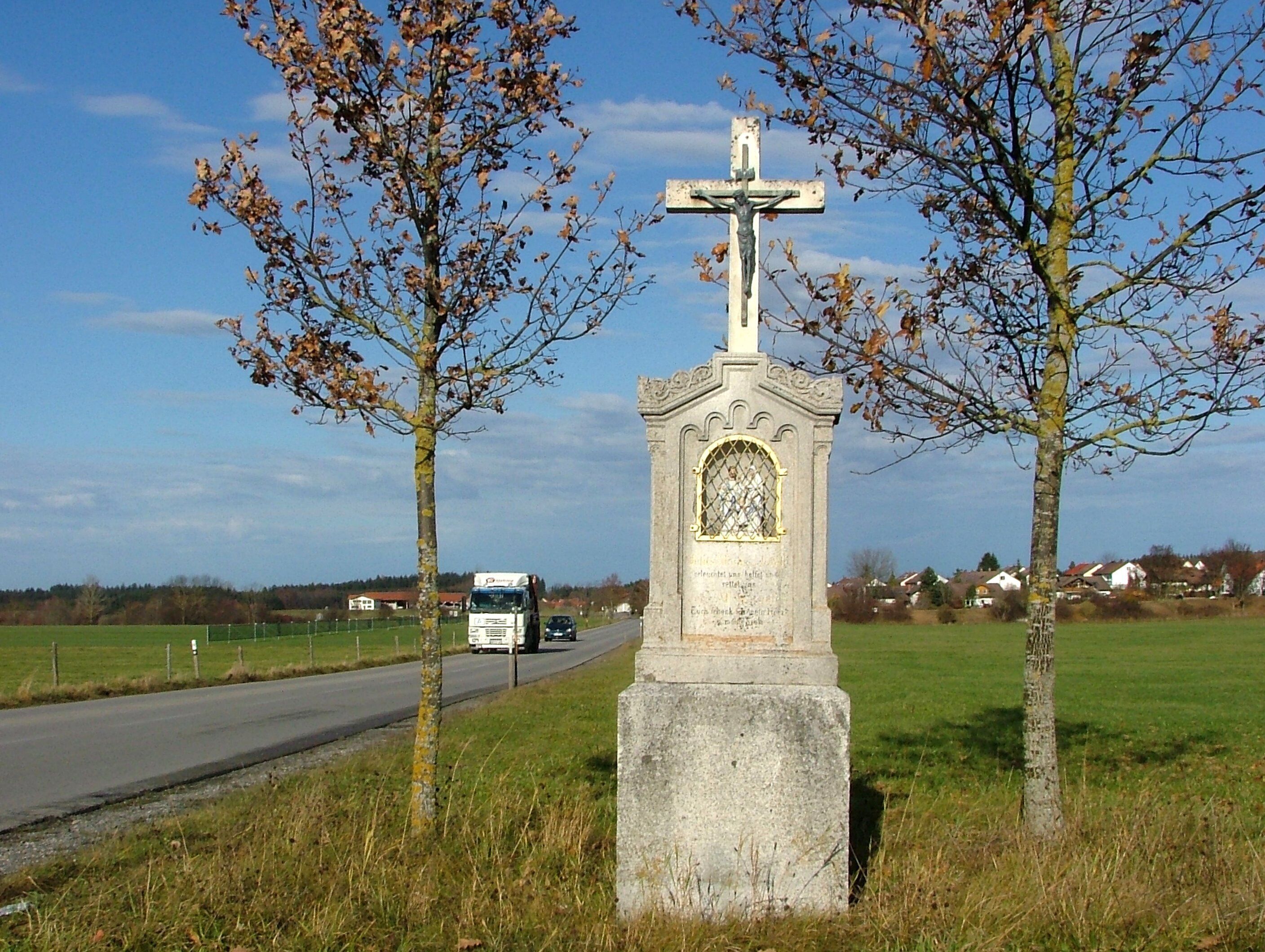 A roadside cross monument with a crucifix, surrounded by trees and a road with a passing truck.