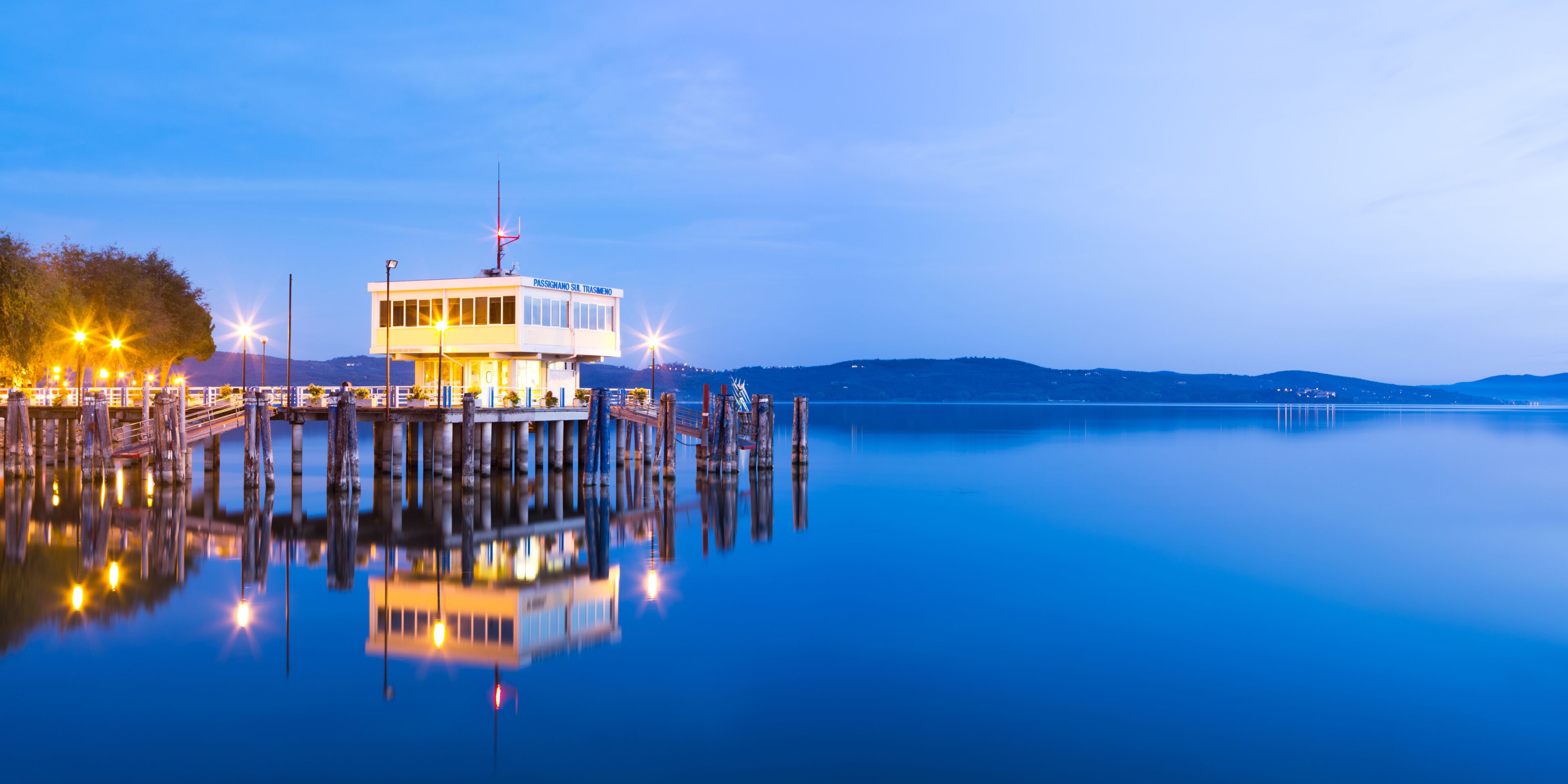 A waterfront building with a dock extending into calm water, surrounded by hills.