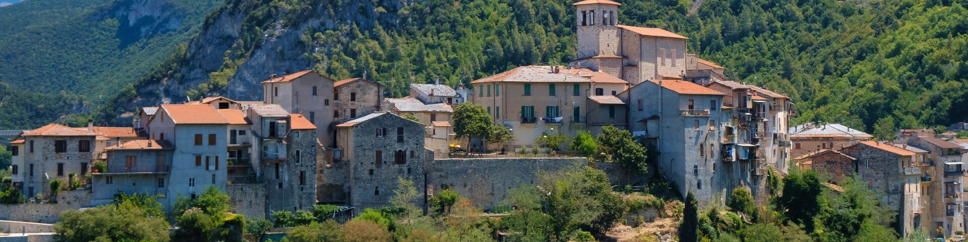 Un pueblo costero con edificios de piedra y una torre destacada, rodeado de vegetación exuberante y montañas.