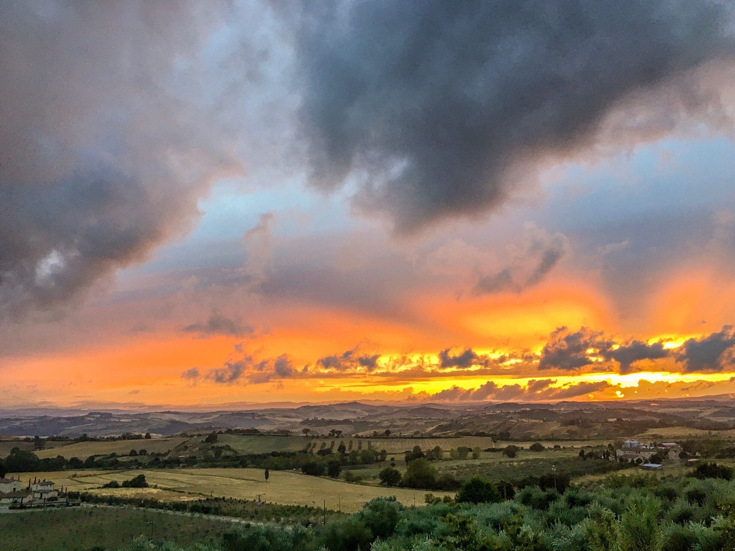 A rural landscape with rolling hills, fields, and scattered buildings under a dramatic sunset sky.