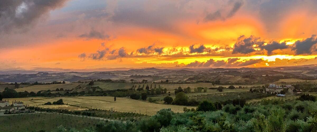 A rural landscape with rolling hills, fields, and scattered buildings under a dramatic sunset sky.