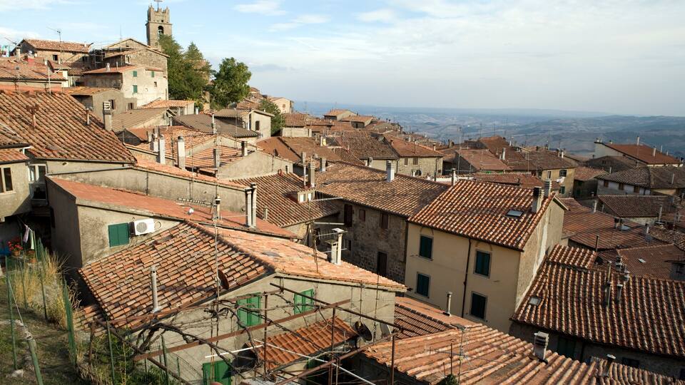 A town with terracotta-tiled rooftops, a prominent church tower, and a hilly landscape in the background.