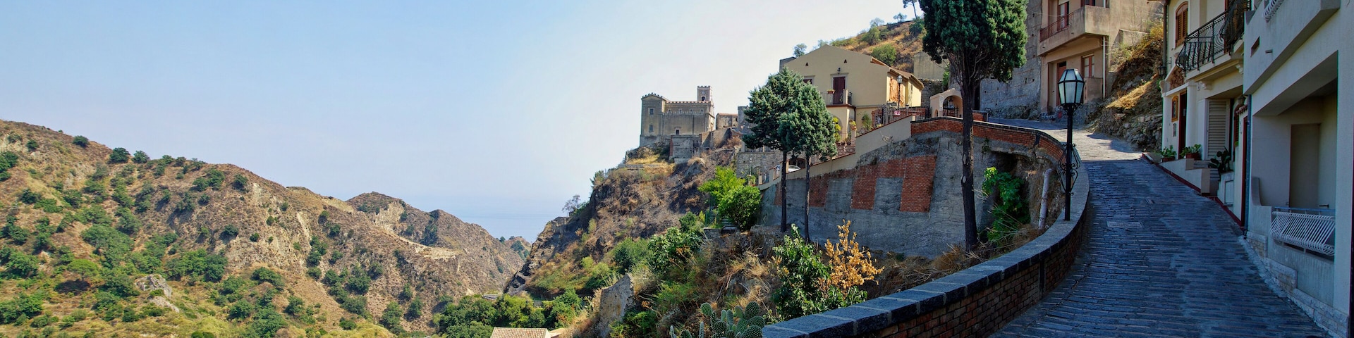 Una calle adoquinada en un pueblo montañoso con edificios, un castillo y cactus.
