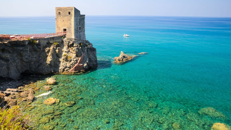 A coastal cliff with a stone tower, clear turquoise waters, and a small boat in the distance.