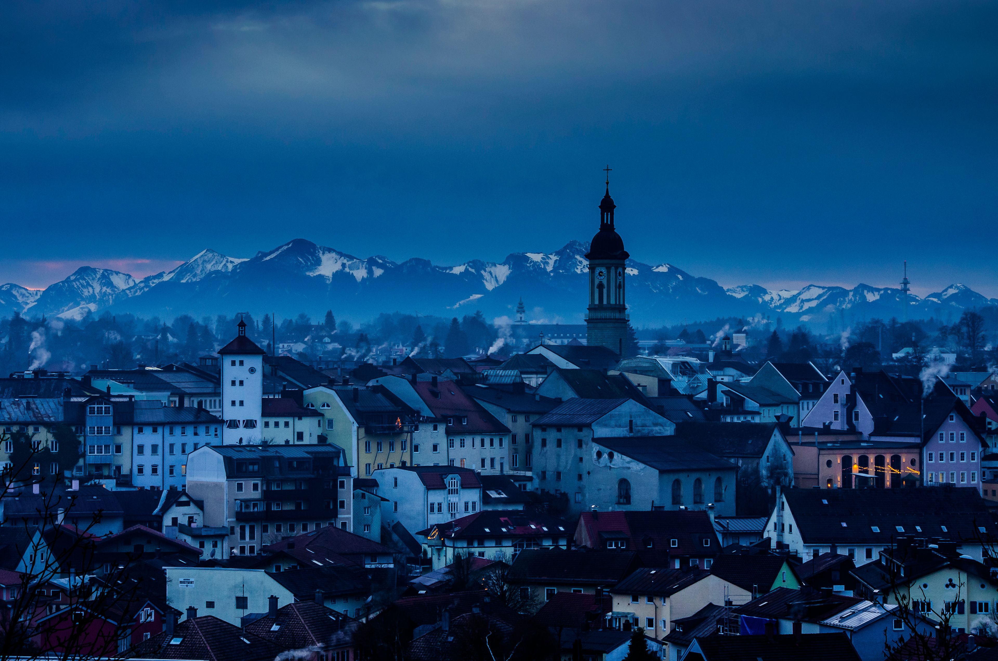 A town with a prominent clock tower, surrounded by snow-capped mountains under a twilight sky.