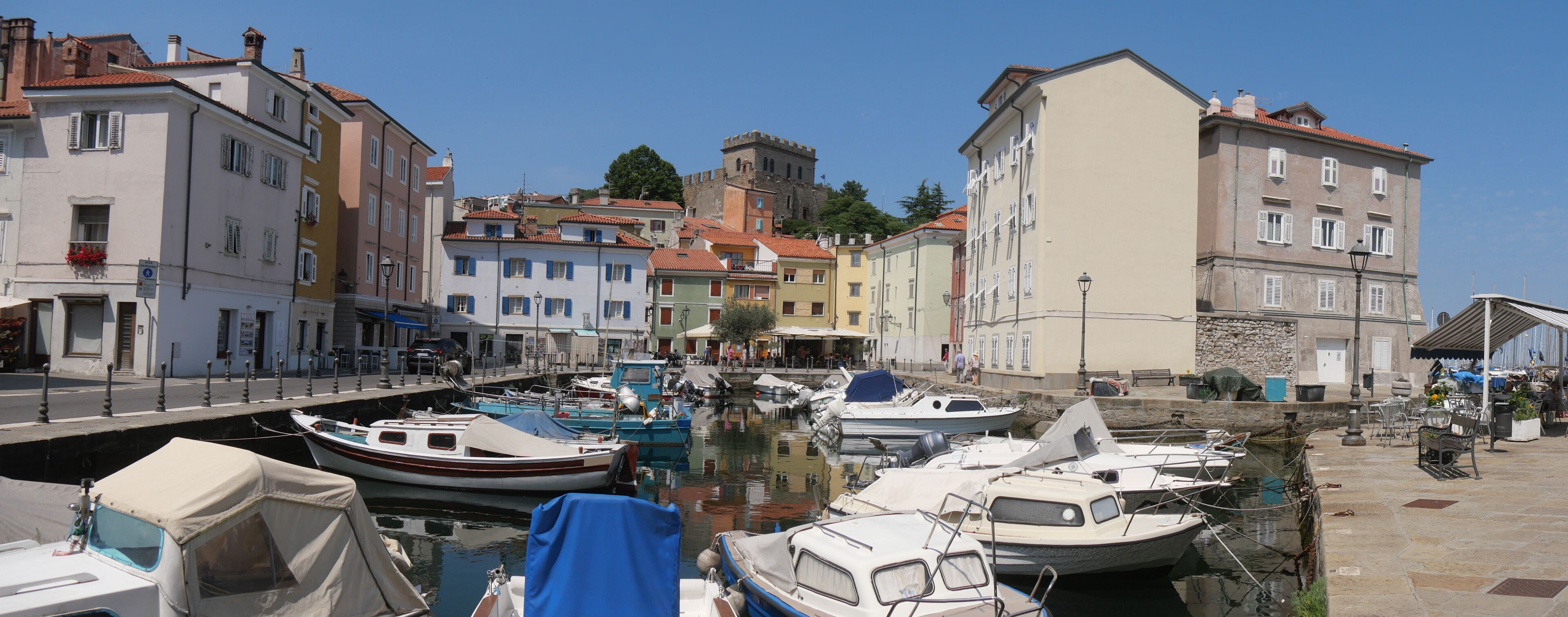 Mandracchio in Muggia : panorama of the small harbor inside the village and surrounded by ancient walls and colorful buildings. There are colored boats moored and reflected in the water.