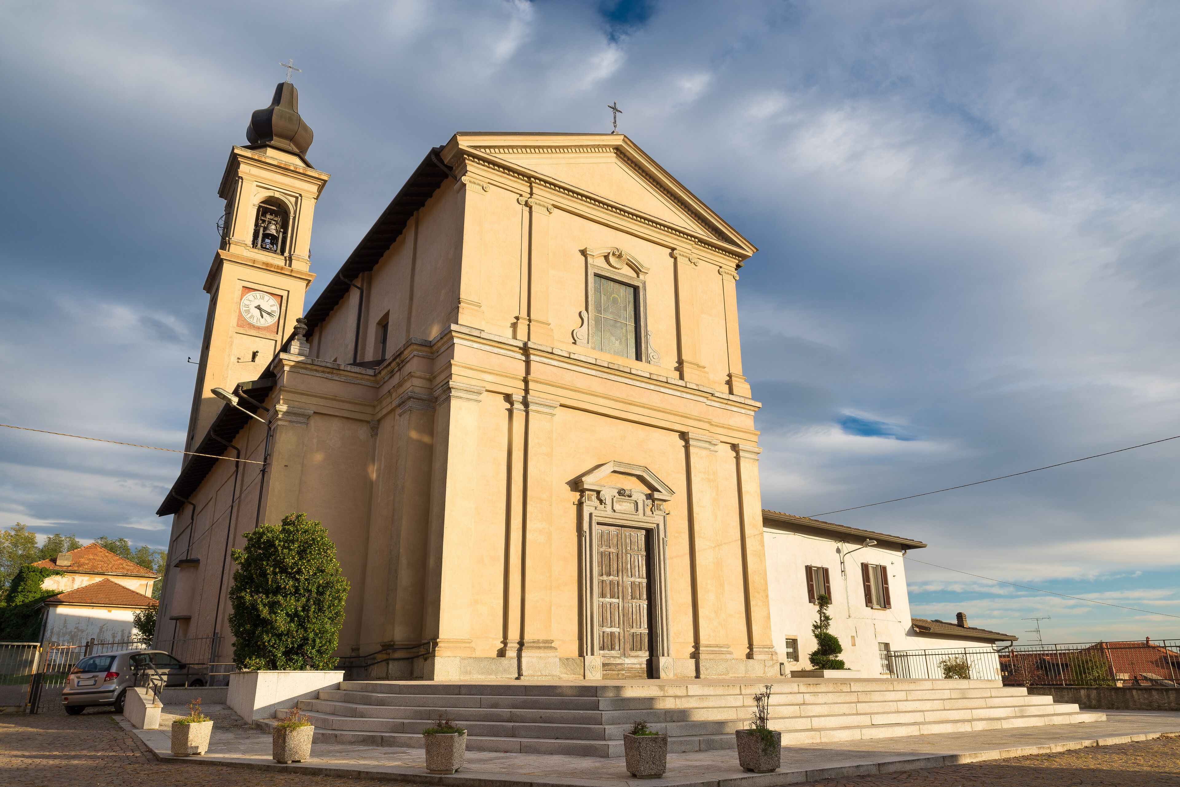 A beige church with a bell tower and a clock, surrounded by steps and potted plants.