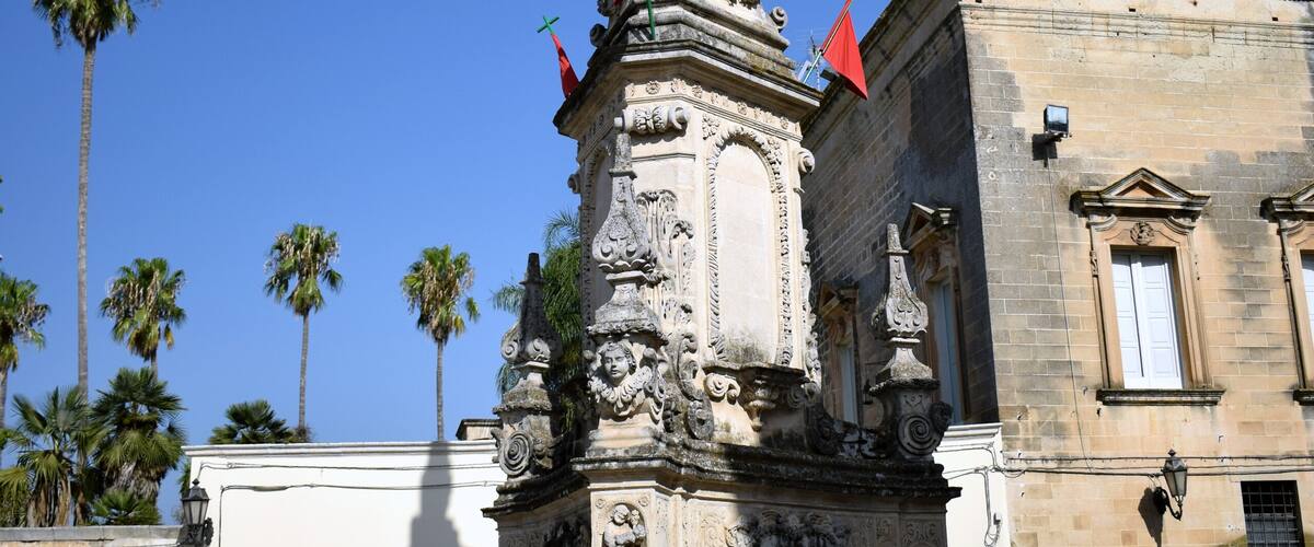 A historical monument with intricate carvings and a red flag on top, surrounded by palm trees and classic architecture.