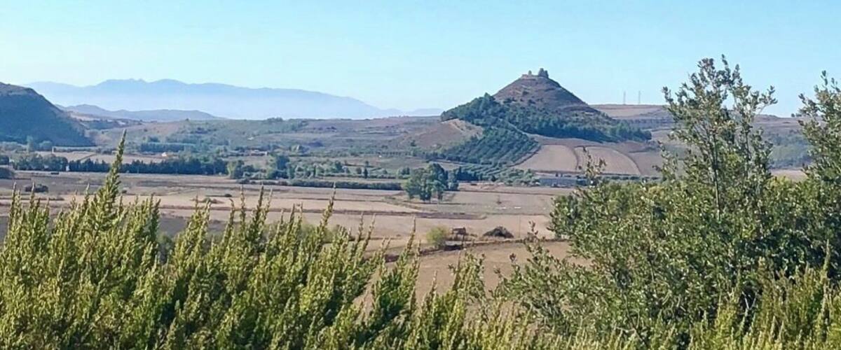 A hilly landscape with a prominent hill in the center and distant mountains under a clear blue sky.