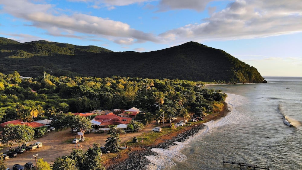 Guadeloupe beach drone sunset view. Plage Caraibe black beach aerial view in Pointe Noire. Backlit ocean wave shadows.