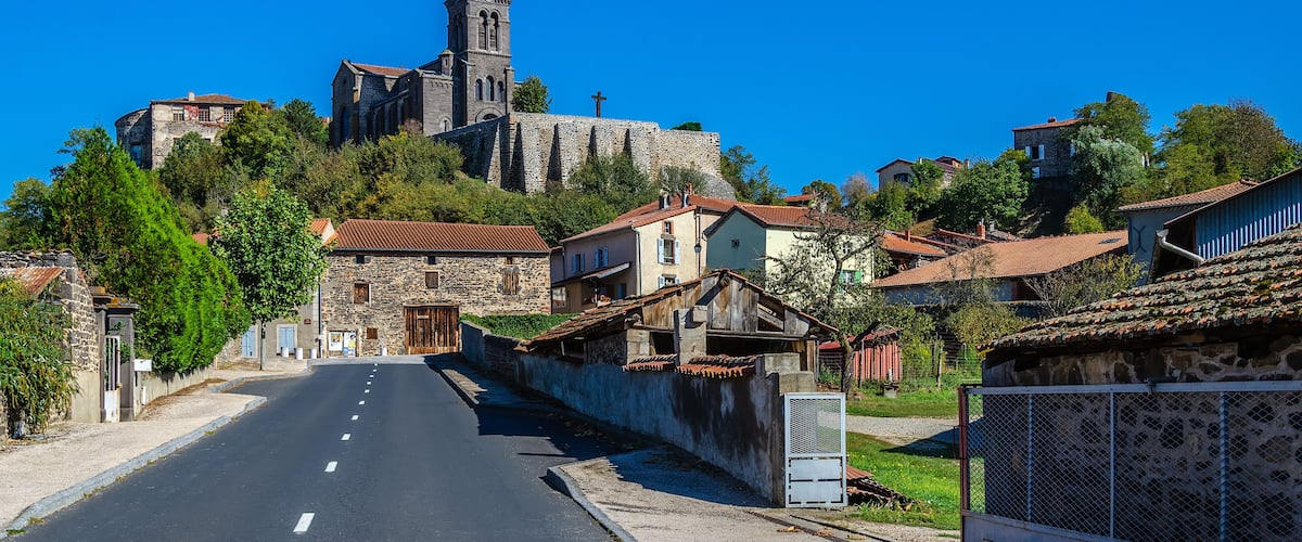 A street leading to a historic church with a tall bell tower, surrounded by traditional stone buildings and lush greenery.