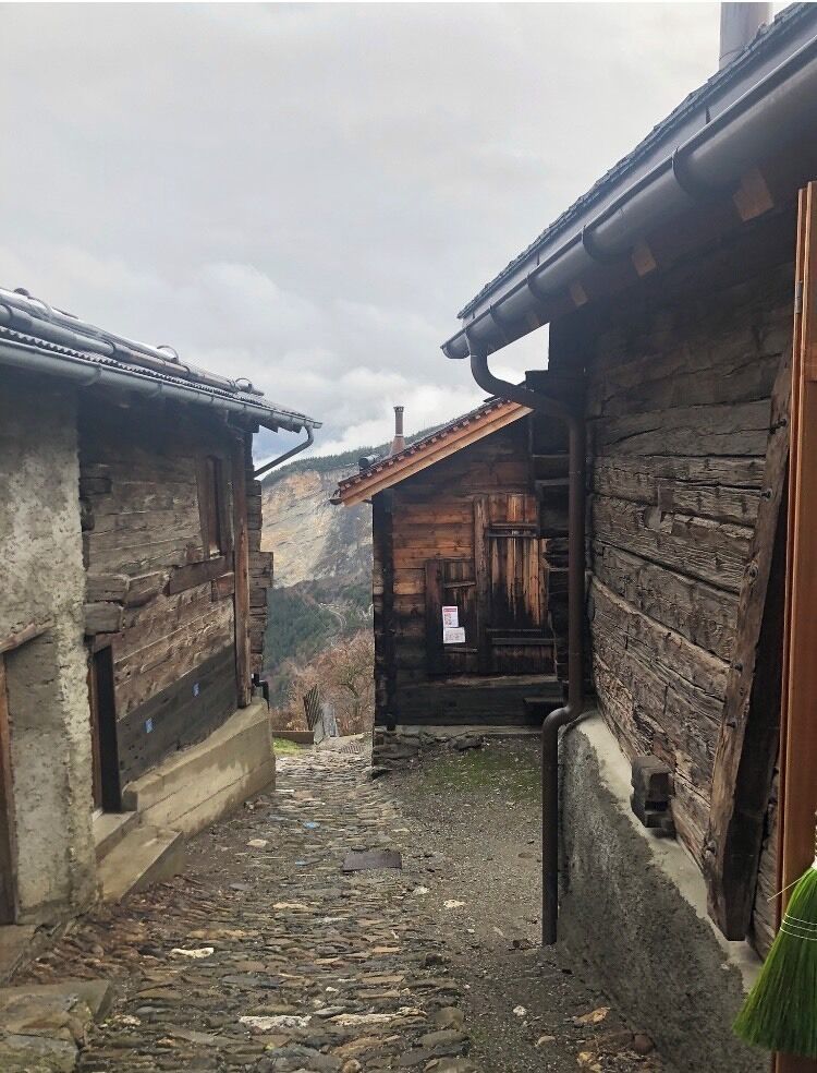 A narrow stone pathway between rustic wooden buildings with a mountainous backdrop.