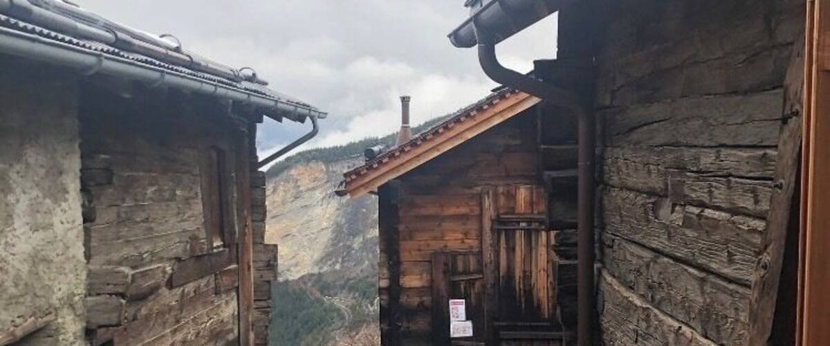 A narrow stone pathway between rustic wooden buildings with a mountainous backdrop.