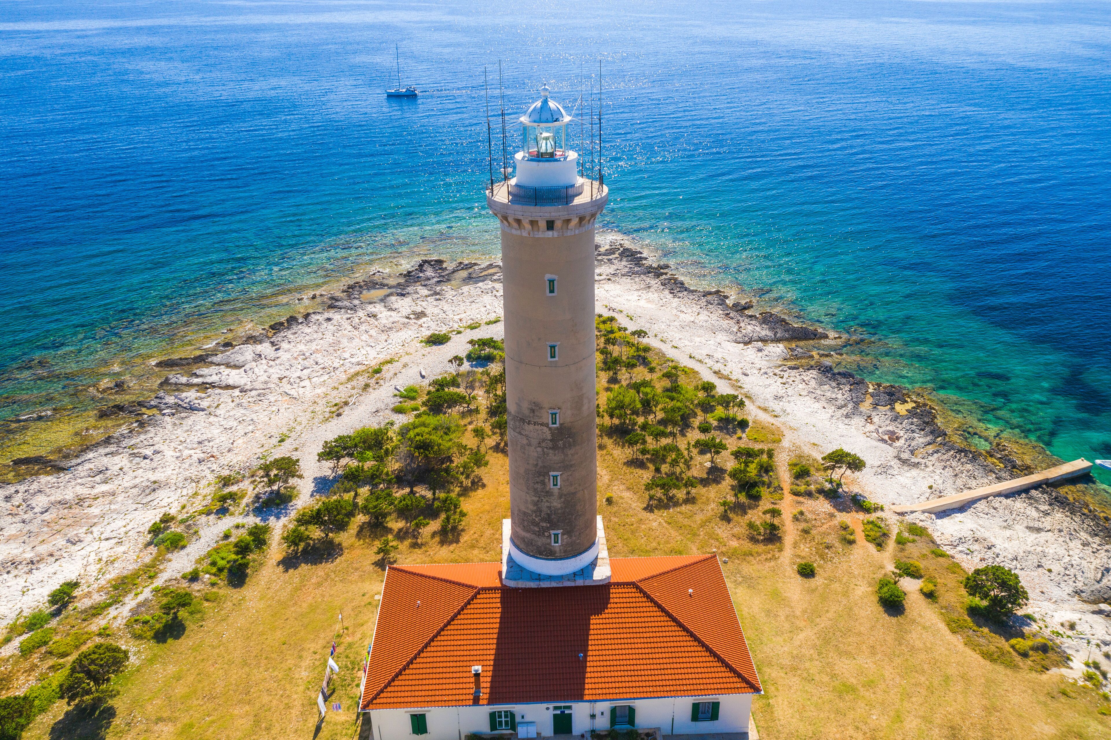 A coastal lighthouse with a red roof, surrounded by clear blue waters and rocky terrain.