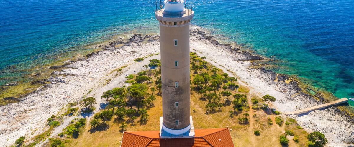A coastal lighthouse with a red roof, surrounded by clear blue waters and rocky terrain.