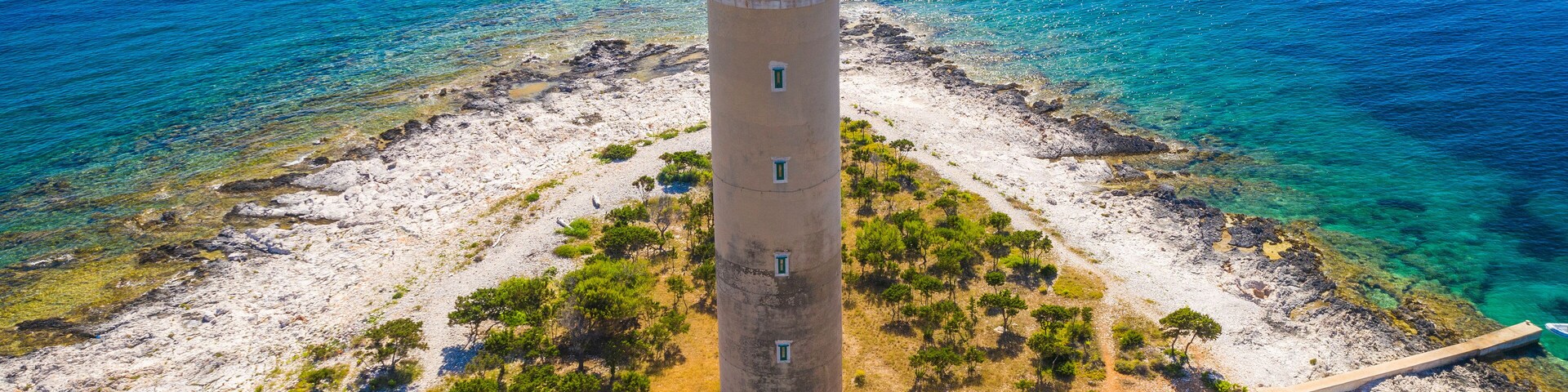 A coastal lighthouse with a red roof, surrounded by clear blue waters and rocky terrain.