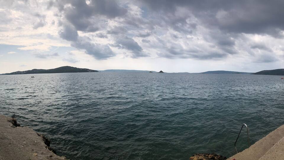 A coastal scene with a stone wall, calm sea, distant islands, and overcast sky.