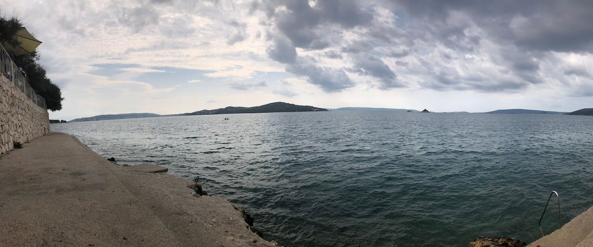 A coastal scene with a stone wall, calm sea, distant islands, and overcast sky.