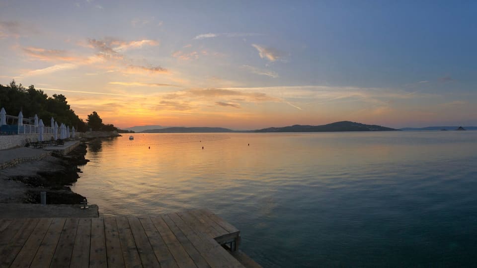 A calm sea at sunset with boats, a wooden pier, and distant hills.