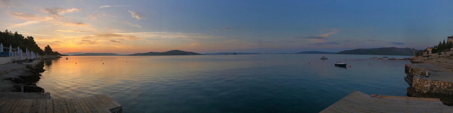 Mar tranquila al atardecer, con barcos, un muelle de madera y colinas a lo lejos.